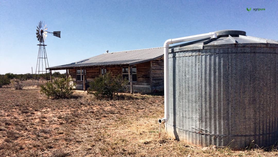 A large rainwater collection tank beside a Texas farmhouse with gutters feeding it, dry land around