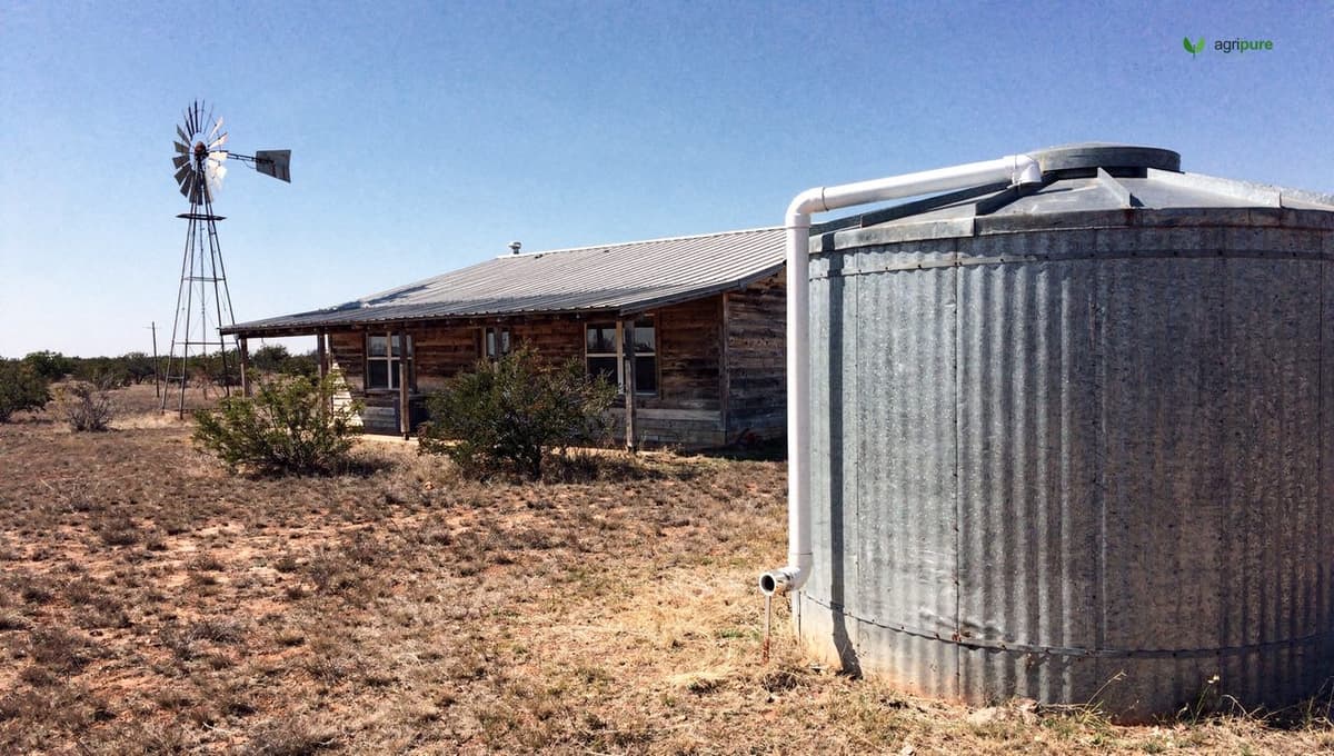 A large rainwater collection tank beside a Texas farmhouse with gutters feeding it, dry land around
