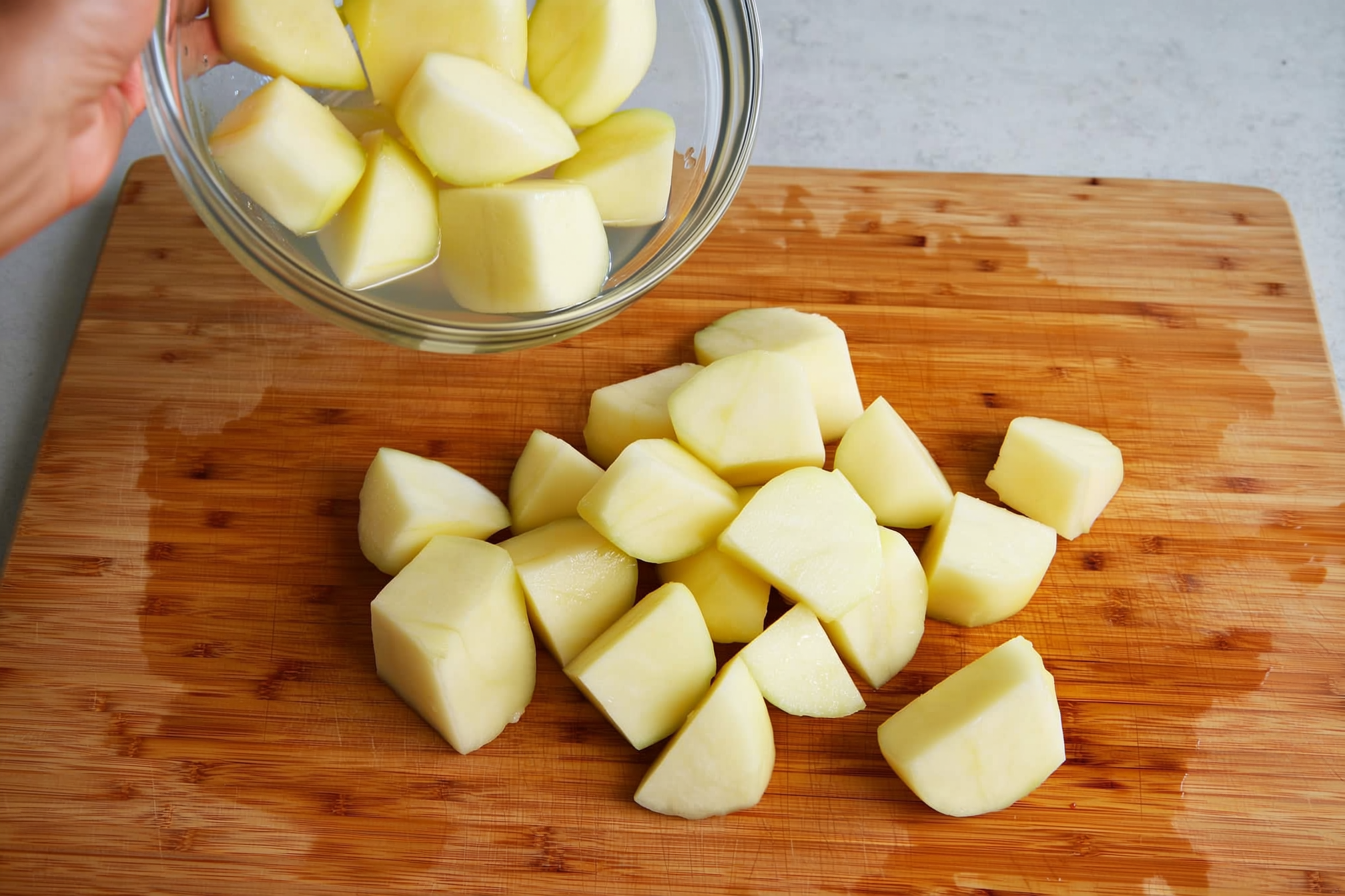Peeled white potatoes being cut into half-inch cubes on a wooden cutting board, with a bowl of ascorbic acid water nearby to drop the cubes into immediately