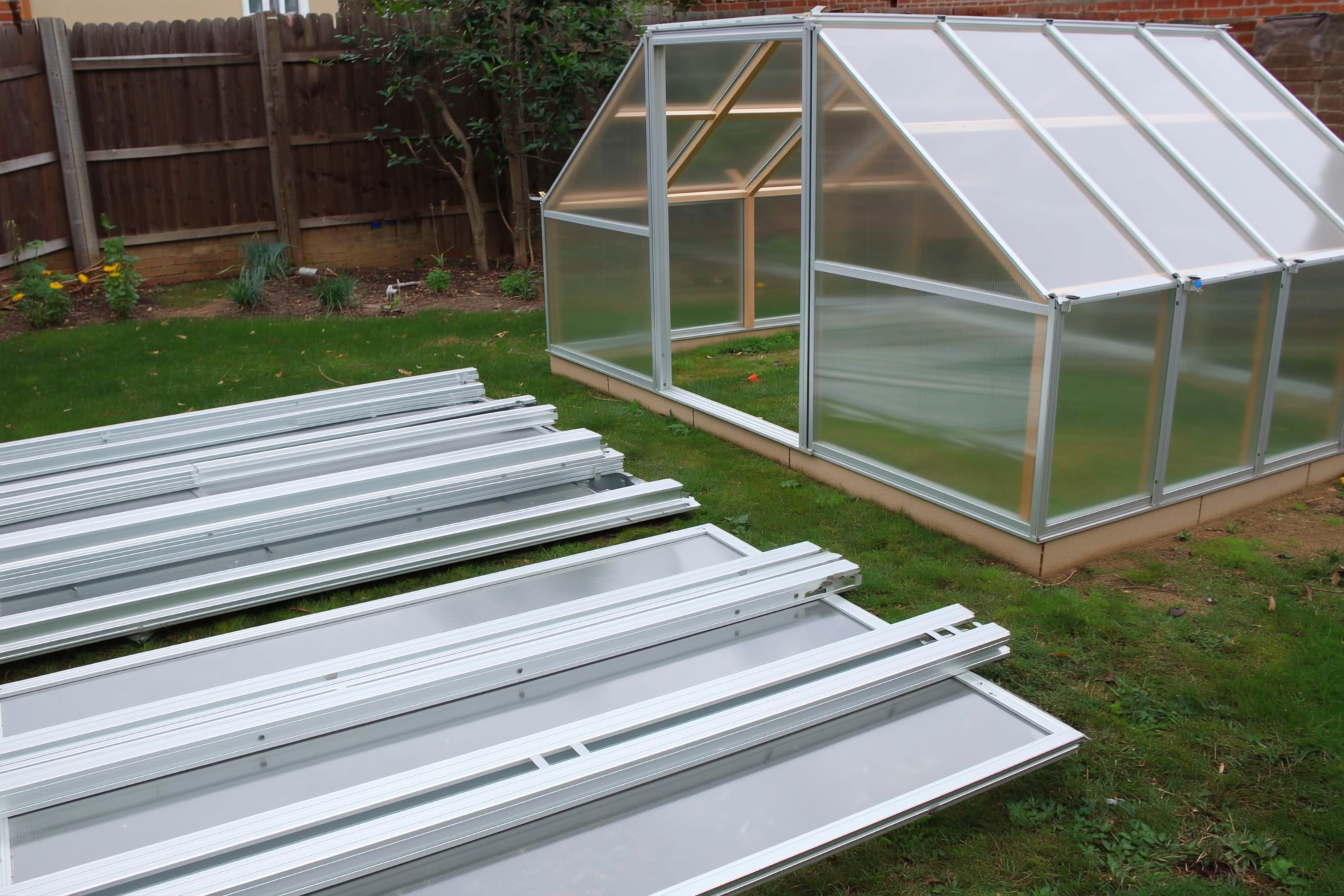 A flat-pack greenhouse kit being assembled in a backyard, aluminum frame parts and polycarbonate panels laid out with a half-built frame standing, no people