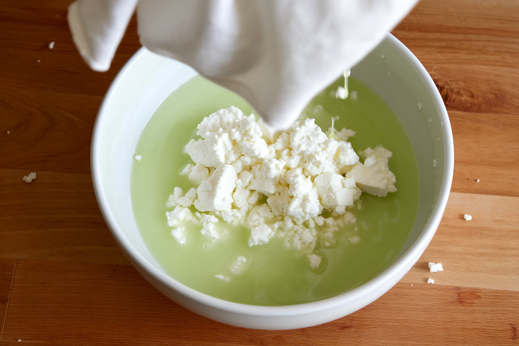 Fresh white cheese curds draining in a cheesecloth-lined colander over a bowl of pale green whey on a wooden counter, the simplest fresh-cheese method in progress