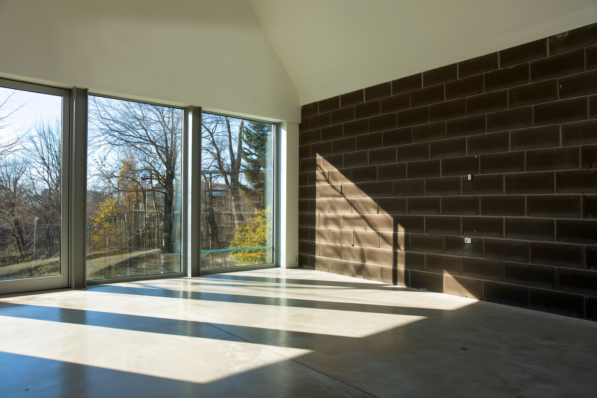 Winter afternoon sun streaming through large south-facing windows onto a polished concrete floor and a dark brick thermal-mass wall inside a passive solar home
