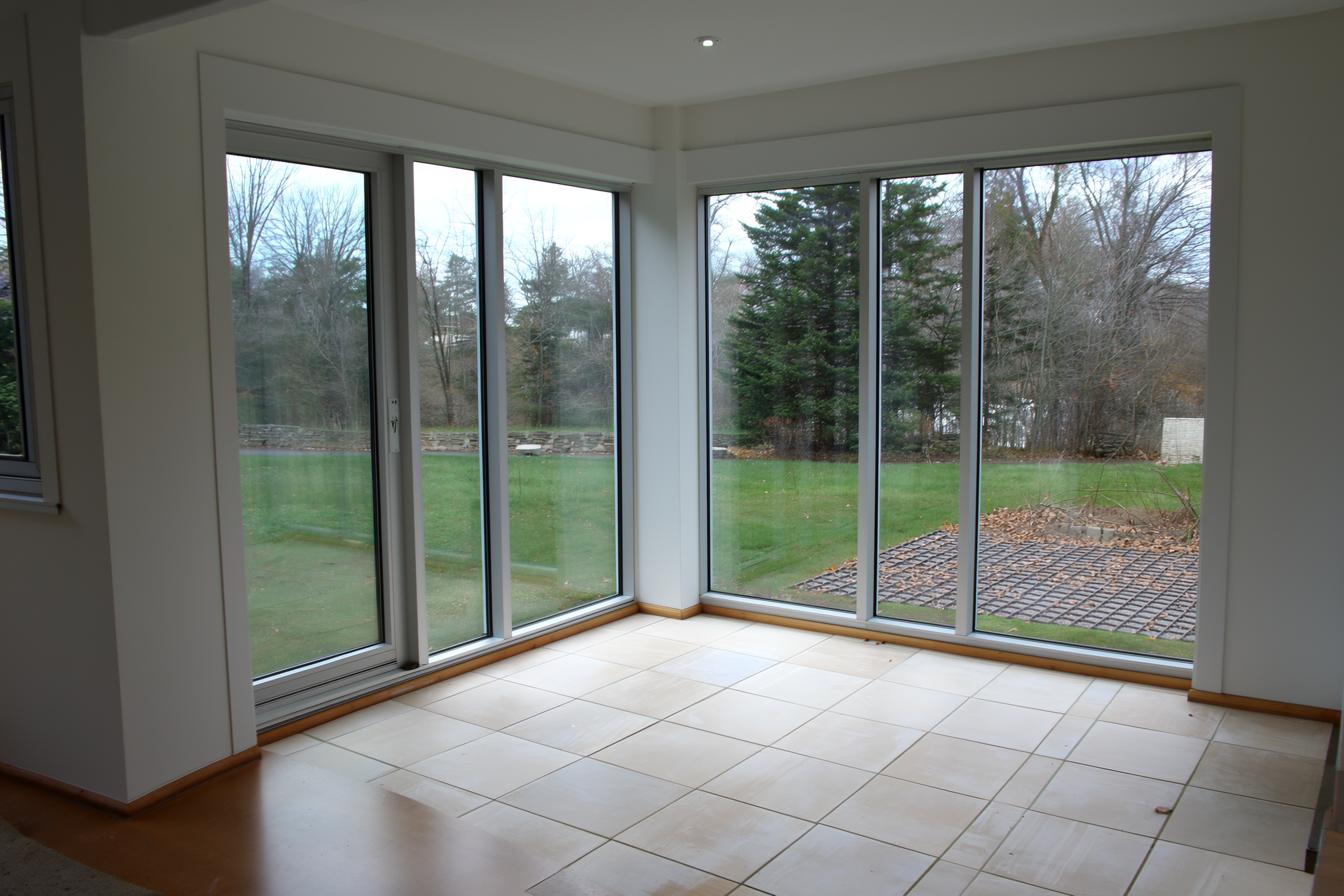 An attached glazed sunspace on the south side of a home with floor-to-ceiling windows and a tiled thermal-mass floor warmed by low winter sun, connected to the house by a glass door