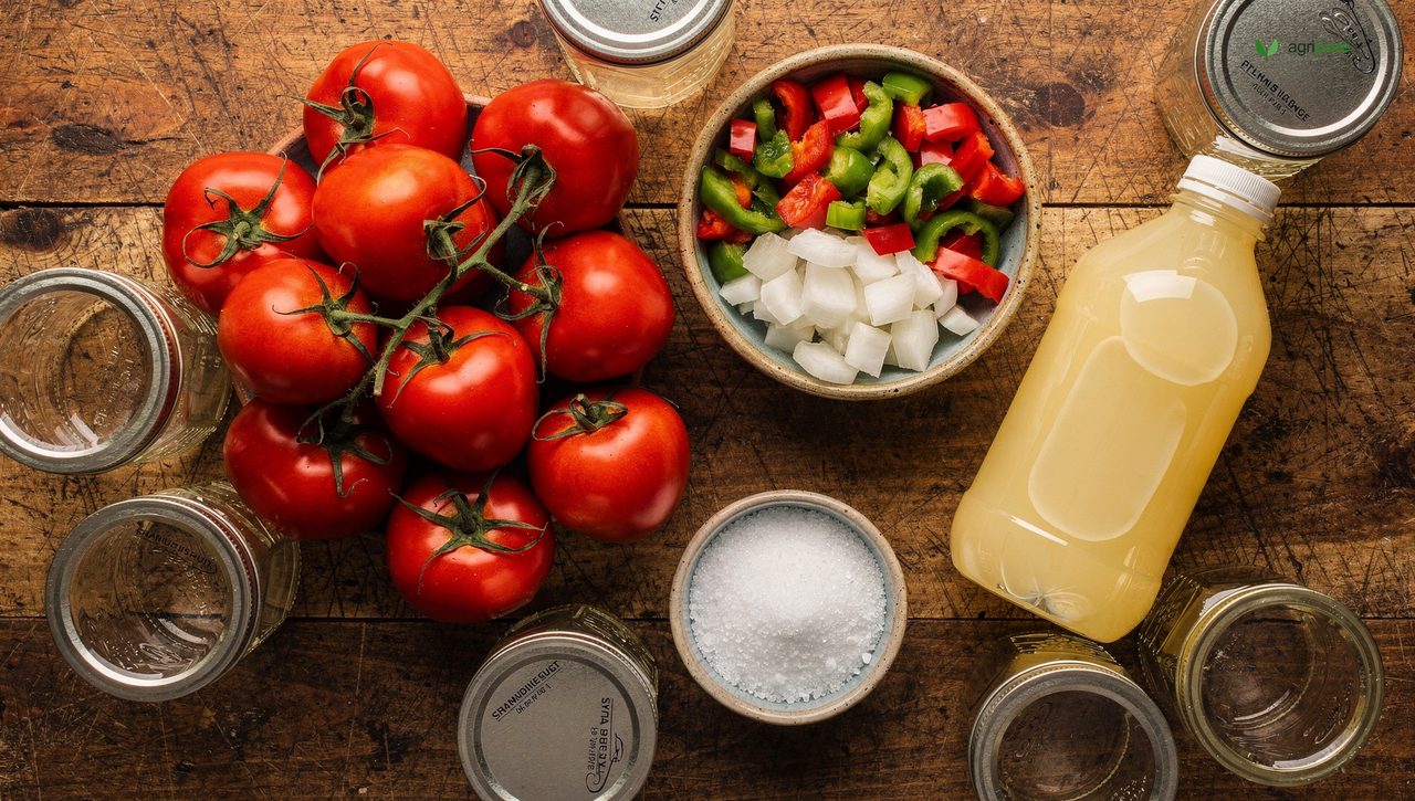 Overhead shot of fresh salsa ingredients laid out for canning — a pile of ripe red tomatoes, bowls of diced onion and mixed peppers, a bottle of lemon juice, and canning salt on a wooden surface