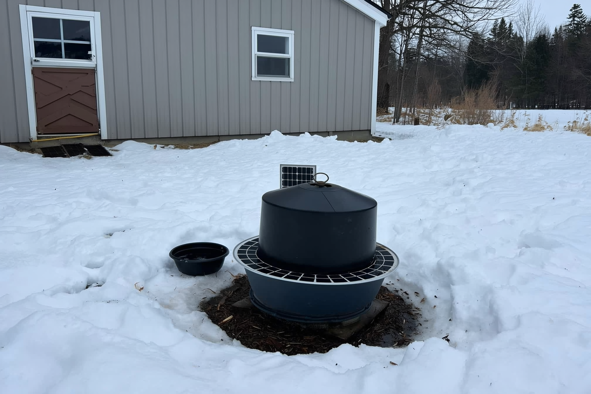A heated poultry waterer and a small dark water bowl in a snowy chicken run beside a coop, a compact solar panel on the coop roof, winter morning, no people