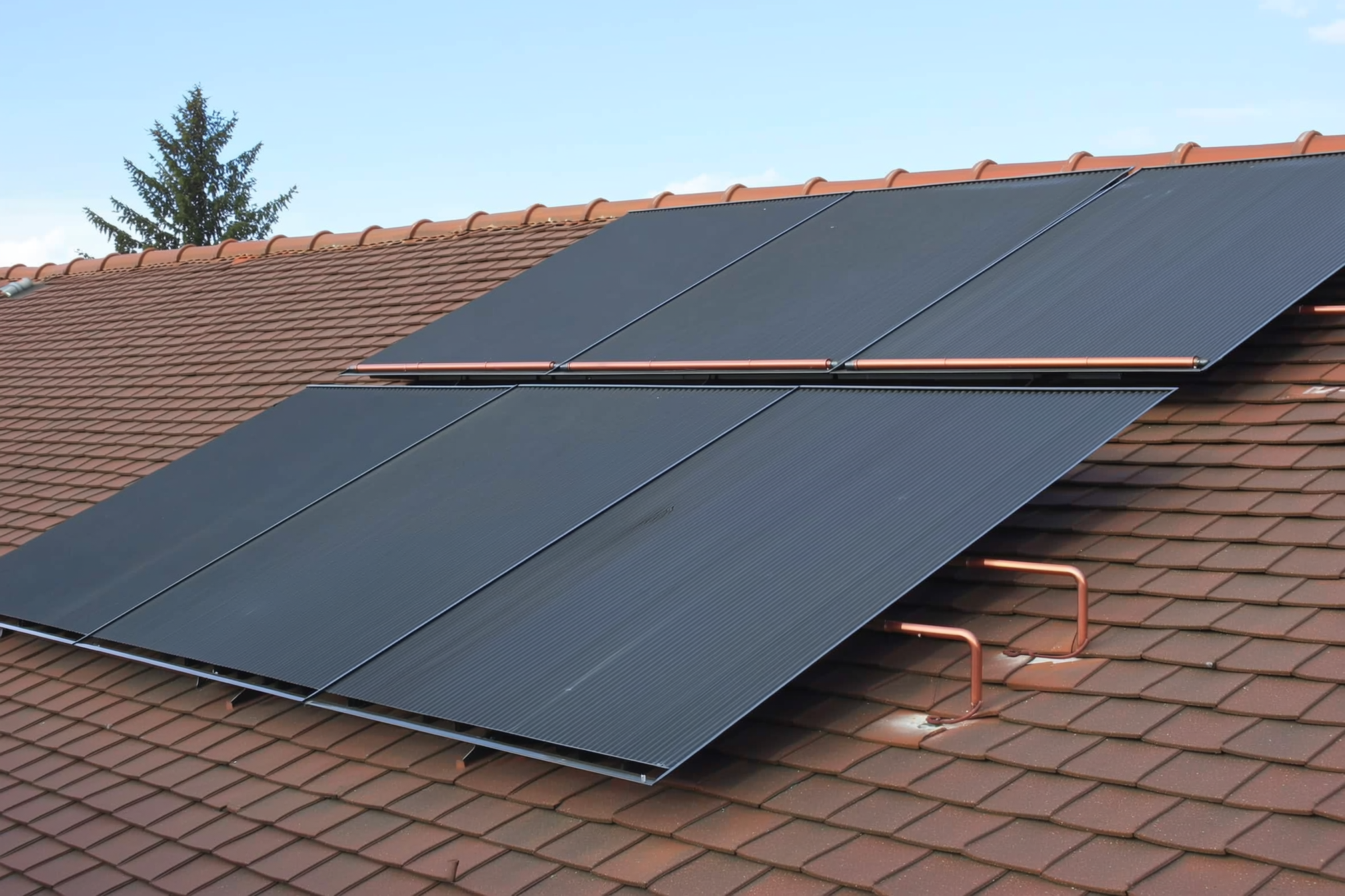 A rooftop solar water heating collector array on a residential roof, dark flat-plate panels angled toward the sun with copper plumbing headers, no people